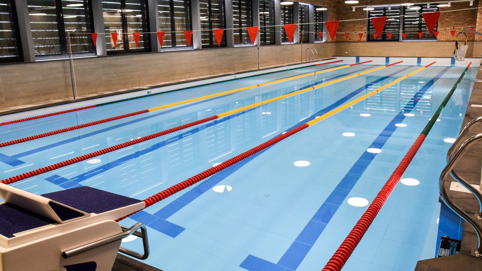Kuldīgas peldbaseins. Iekštelpu peldbaseins ar gaiši zilu ūdeni, vairākām zilām celiņu līnijām un sarkani dzeltenām celiņu virvēm. Priekšplānā redzams starta bloks ar numuru "1", bet virs baseina izkārtoti sarkani karodziņi.