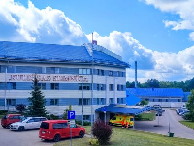 Kuldīgas slimnīcas galvenā ēka ar ziliem jumtiem un gaišām fasādēm. Uz ēkas ir uzraksts "KULDĪGAS SLIMNĪCA".