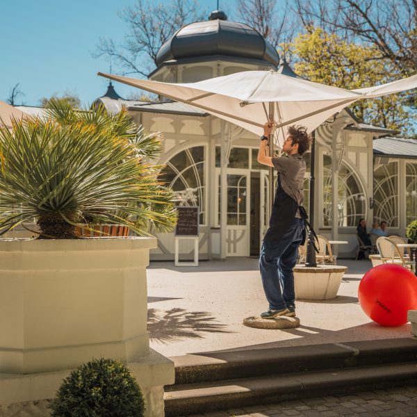 Vīrietis pelēkā kreklā un melnā priekšautā uzstāda lielu, gaišu saulessargu uz saulainas āra terases. Aiz viņa redzama eleganta ēka ar arkveida logiem un kupolu. Terasi rotā podos stādīti augi un liela sarkana piepūšamā bumba.