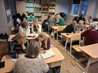 Sievietes sēž pie skolas galdiem klasē, iesaistoties diskusijās un pētot materiālus apmācību sesijā.