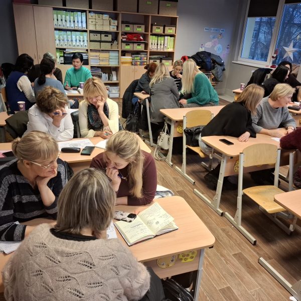 Sievietes sēž pie skolas galdiem klasē, iesaistoties diskusijās un pētot materiālus apmācību sesijā.