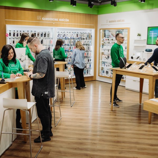 Moderns elektronikas veikals, kurā darbinieki zaļos džemperos apkalpo klientus.