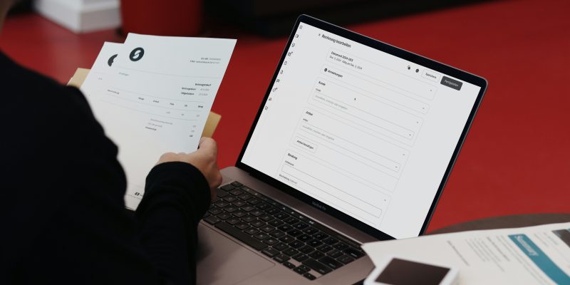 Persona salīdzina papīra rēķinu ar portatīvā datora ekrānā atvērtu programmu.