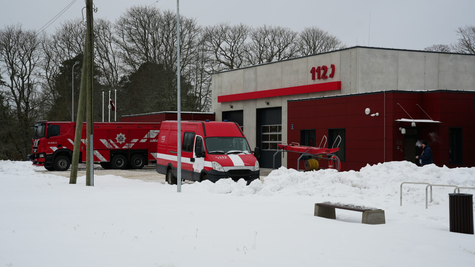 Sarkani-pelēka ugunsdzēsēju depo ēka ar uzrakstu "112" uz fasādes, ietverta sniegā. Pie tās stāv liela sarkana ugunsdzēsēju automašīna ar uzrakstu "OLAINES" un mazs sarkani balts mikroautobuss ar "112" un "2412" uz sāniem. Cilvēks redzams pie ēkas ieejas, fonā kaili koki.