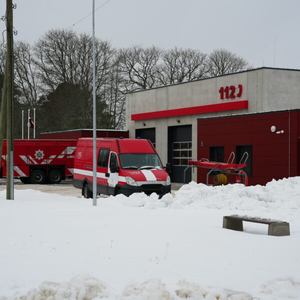 Sarkani-pelēka ugunsdzēsēju depo ēka ar uzrakstu "112" uz fasādes, ietverta sniegā. Pie tās stāv liela sarkana ugunsdzēsēju automašīna ar uzrakstu "OLAINES" un mazs sarkani balts mikroautobuss ar "112" un "2412" uz sāniem. Cilvēks redzams pie ēkas ieejas, fonā kaili koki.