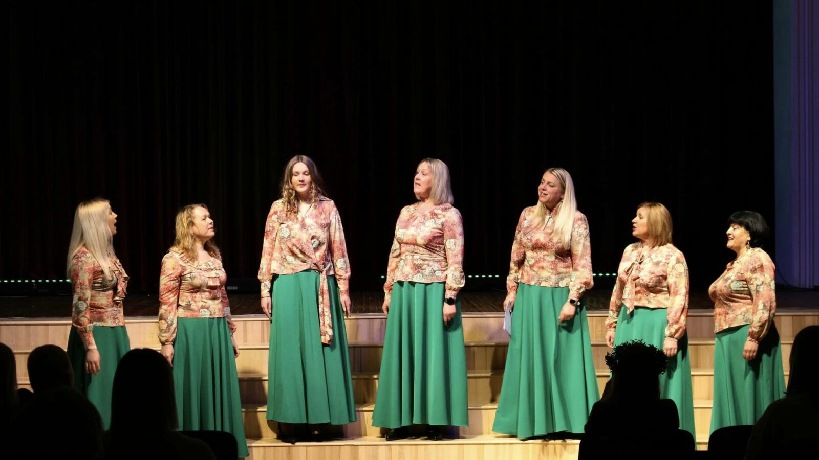 Septiņas sievietes saskaņotos tērpos – ziedu raksta blūzēs un garos, zaļos svārkos – uzstājas uz skatuves. Viņas stāv rindā pret tumšu fonu, dažas skatās viena uz otru, dažas uz priekšu, un viena sieviete dzied ar atvērtu muti. Priekšplānā redzami skatītāju silueti.