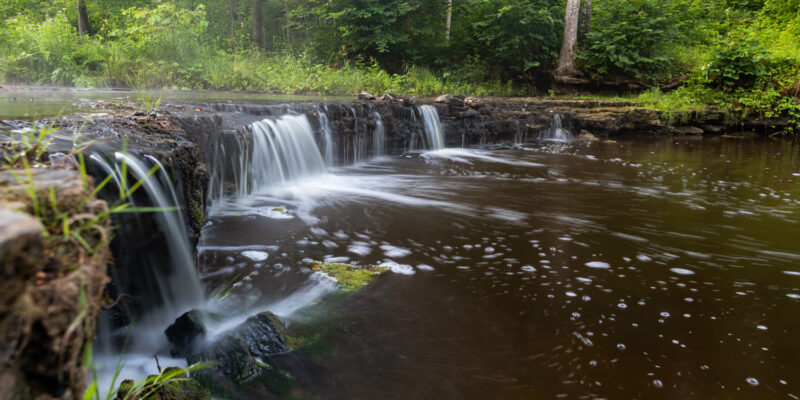 Ūdenskritums ar plūstošu, baltu ūdeni, kas krīt pāri tumšiem akmeņiem brūnganā upē, ko ieskauj zaļš mežs.